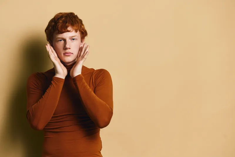 Young man with red hair wearing brown turtleneck posing with hands near face on beige background for portrait and fashion concept.
