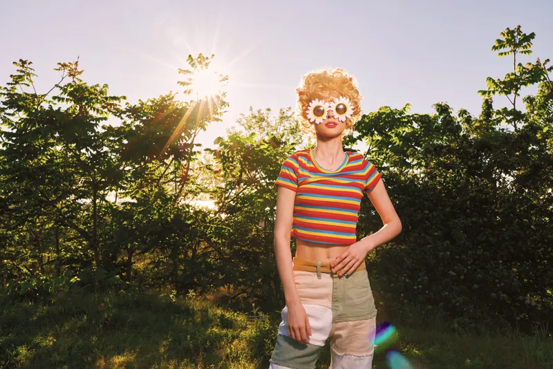 A young woman with blonde curly hair wearing a striped crop top and shorts stands in a sunlit field, wearing a floral mask and projecting a playful retro vibe.
