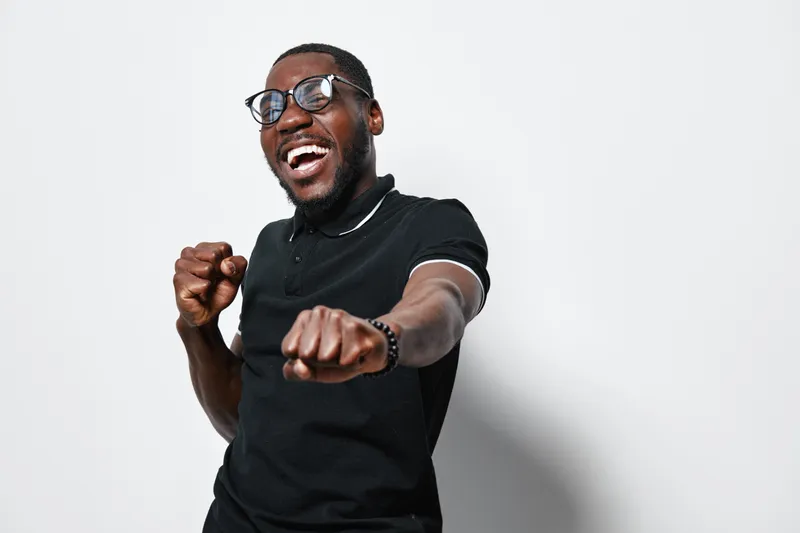 African American man with dark skin and glasses smiling and posing with fists forward in black t shirt against white background for happiness and confidence.