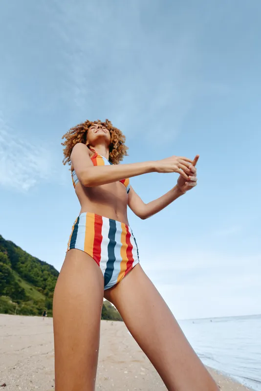 Woman in colorful swimsuit stretching arms on sandy beach by ocean under blue sky, curly hair blowing, summer vacation mood, fitness and wellness outdoors, in warm sunlight and fresh sea air