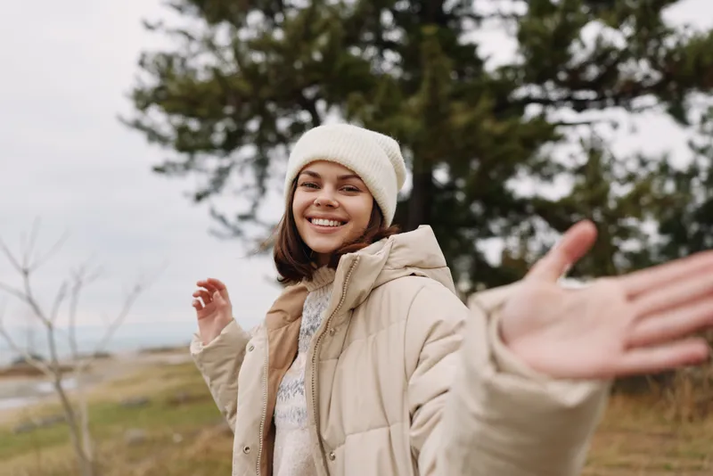 A woman outdoors with a warm coat and beanie smiles with arms open, embracing winter nature and a wide landscape in a peaceful moment