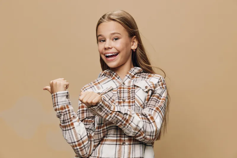Girl smiling and pointing thumb sideways wearing plaid shirt with long hair and natural makeup on beige background in studio portrait.