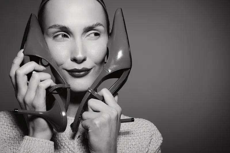 Smiling white woman with light skin holds high heel shoes near face, wears sweater, isolated on grey background. Concept of fashion and style.