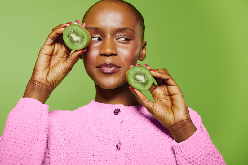 Portrait of a dark skinned African woman holding two kiwi halves near her face. She wears a pink sweater and poses against a green background. Concept of healthy lifestyle and fresh fruit.