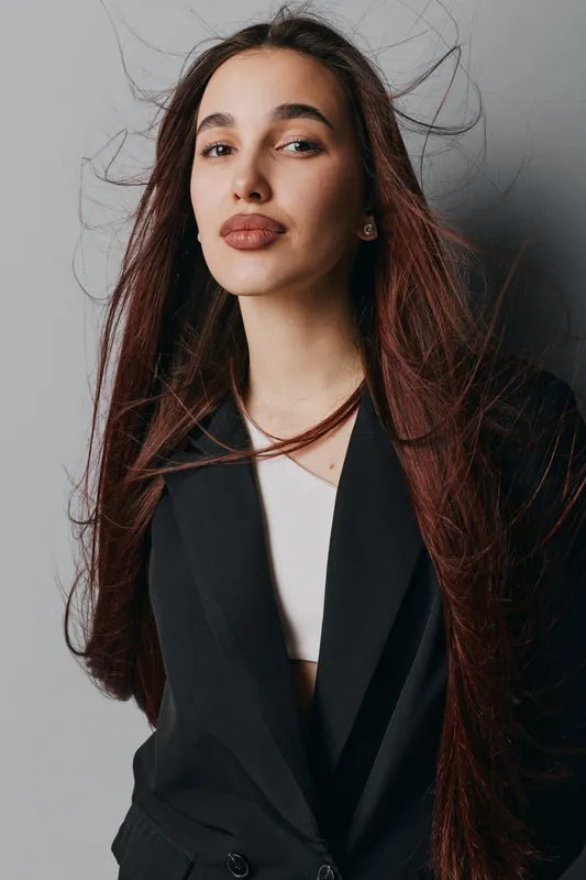 Young woman with long brown hair wearing black blazer and white top posing in studio with confident expression and natural makeup against gray background with hair slightly moving.