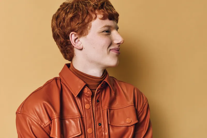 Young man with red hair wearing brown leather jacket and turtleneck smiling looking to side on beige background in studio portrait.