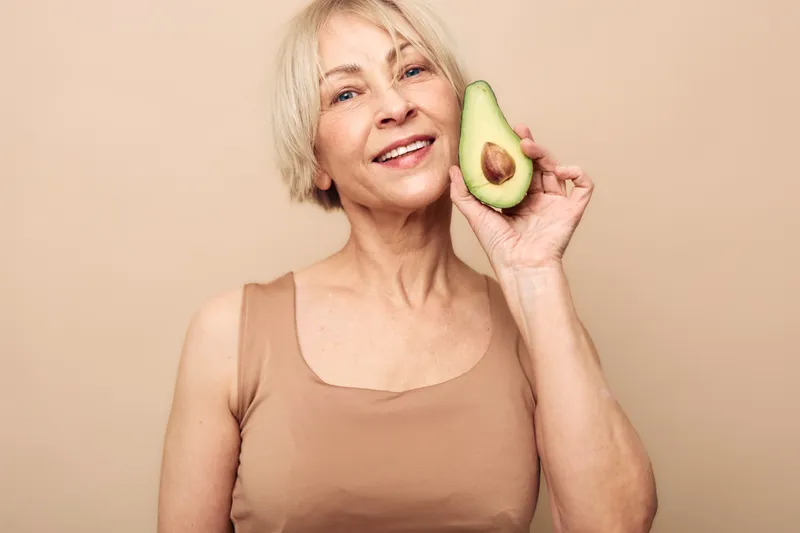 Smiling mature woman with short blonde hair holds half of fresh avocado near face on beige background. Concept of healthy nutrition, natural beauty, wellness, and positive lifestyle for seniors.