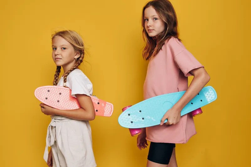 Two girls holding colorful skateboards stand against a yellow background. They wear casual clothes and look confident with relaxed postures.