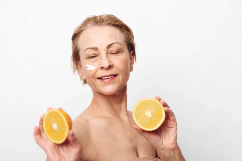 Smiling mature woman with natural skin holds two halves of fresh orange, applying moisturizing cream on face. Concept of healthy skincare, natural beauty, and organic wellness in studio light.