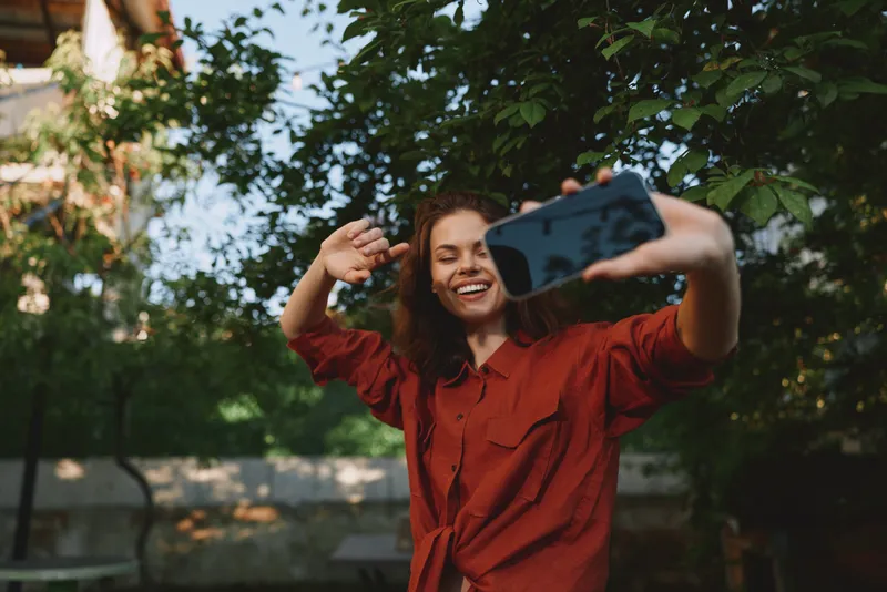 Cheerful woman outdoors, smiling while taking a selfie, red shirt, sunlit moment, relaxed posture, lush greenery background, casual fashion, natural colors, vibrant textures, candid moment