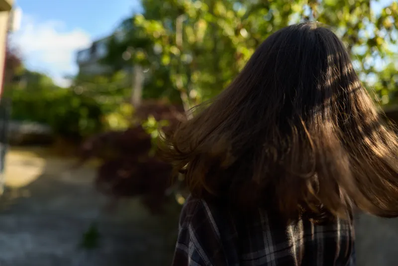 A young woman with long brunette hair wearing a plaid shirt walks outdoors in bright sunlight, captured in a candid lifestyle moment with leaves glowing in the breeze