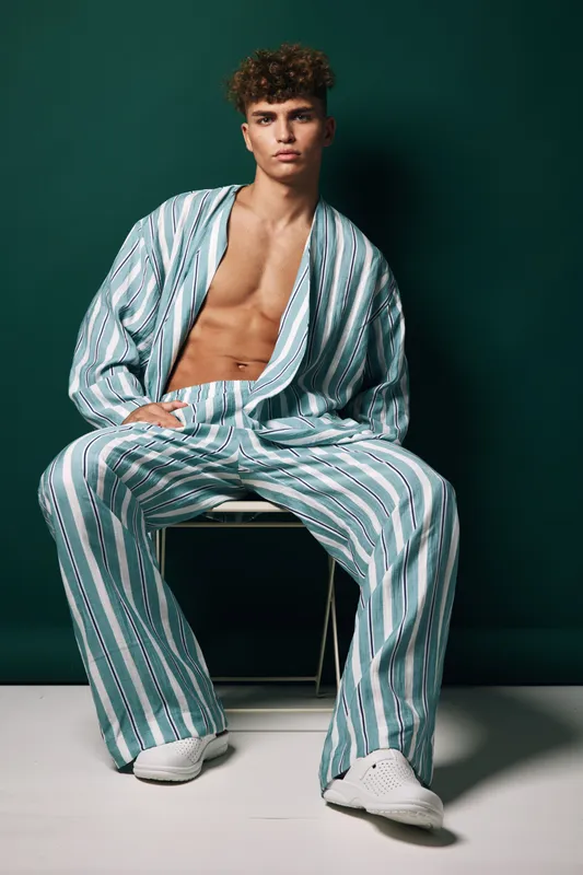 Young man with curly hair wearing striped loose pants and matching open shirt sitting on chair against dark green background with serious expression and white sneakers.