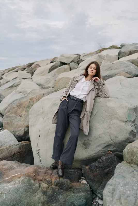 Young woman in casual outfit leans on large rock at rocky shore under cloudy sky. Outdoor fashion portrait with natural landscape and relaxed mood, concept of nature and style.