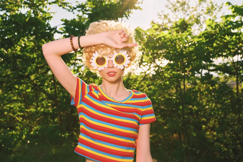 Young woman outdoors wearing a rainbow striped shirt and sunflower sunglasses. Bright natural light creates a playful summer vibe with a retro touch and a carefree lifestyle.