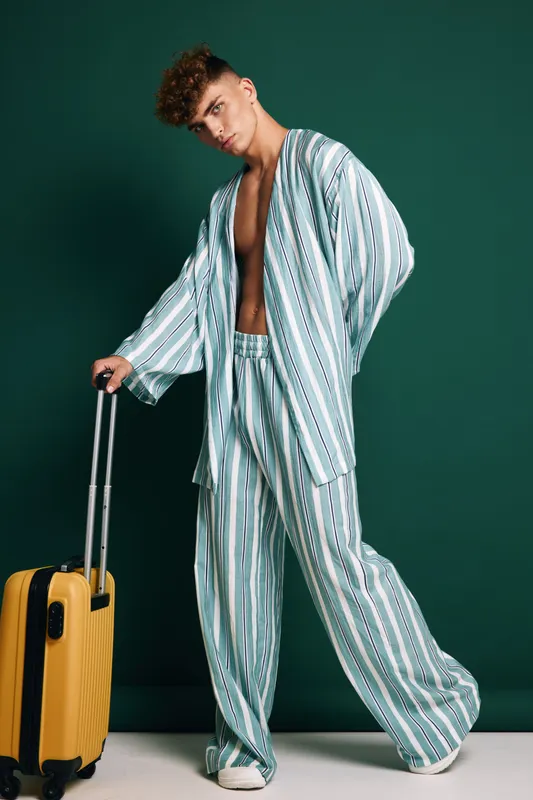 Young man in striped pajamas standing with yellow suitcase on green background, casual travel style, relaxed pose and modern fashion for trip or vacation.