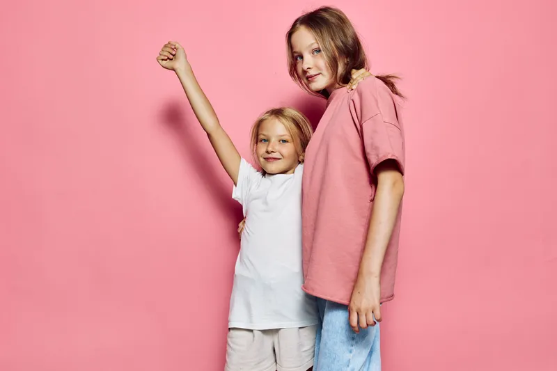 Two smiling girls pose in casual clothes against a pink background, one girl raises her arm confidently. Concept of childhood and friendship.