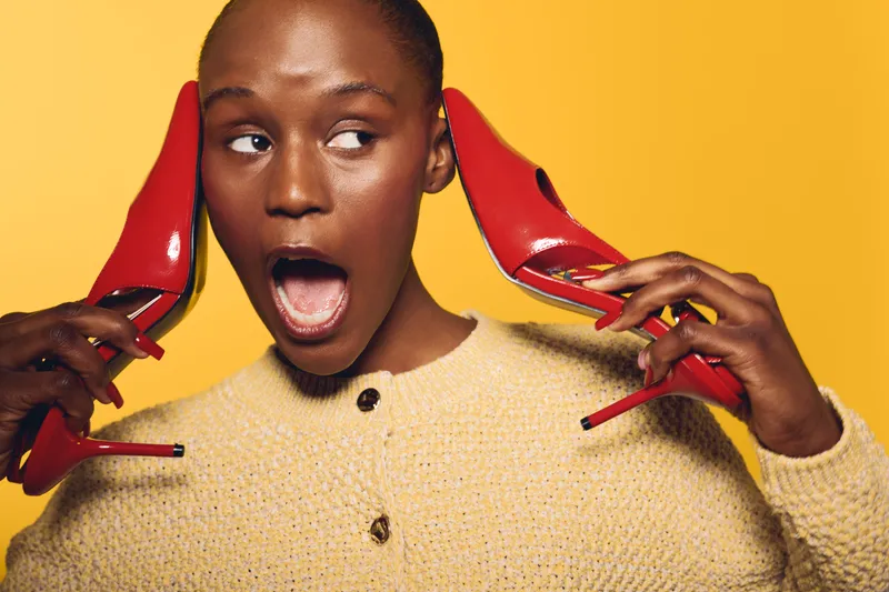 Surprised black woman with dark skin holds red high heels near her ears on yellow background. Concept of fashion, beauty, and playful emotions in studio portrait.