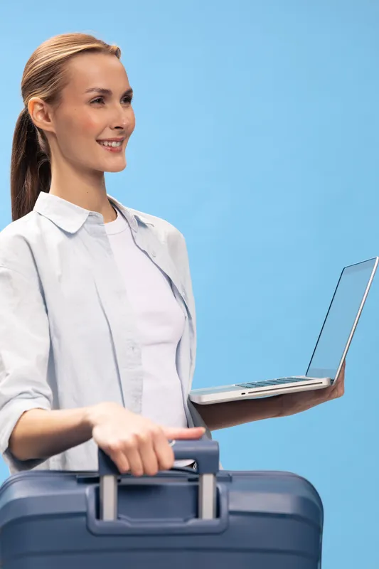 Smiling woman holds suitcase and laptop on blue background. Casual travel concept with technology and business elements. Positive female traveler ready for trip and work.