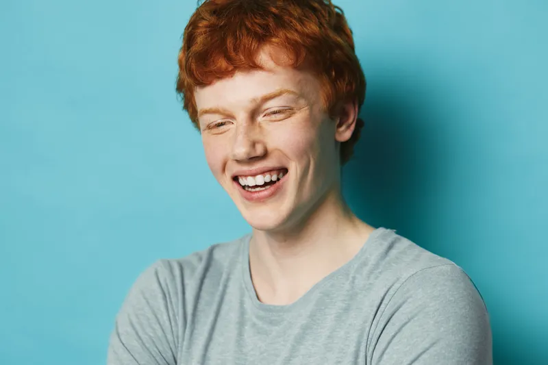 Young man with red hair smiling brightly against blue background, wearing grey shirt. Happy cheerful male portrait with natural light and casual clothing in studio setting.