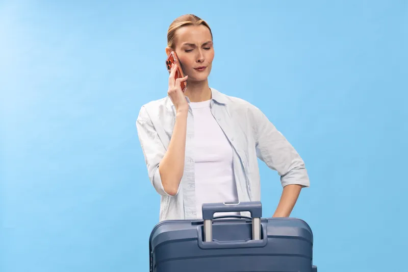 Young woman wearing casual clothes talking on smartphone while standing with blue suitcase on blue background. Concept of travel planning, communication, and vacation preparation.