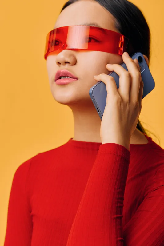 Woman with red futuristic sunglasses talking on smartphone in red sweater on orange background. Modern technology, communication and style with digital device and casual fashion.