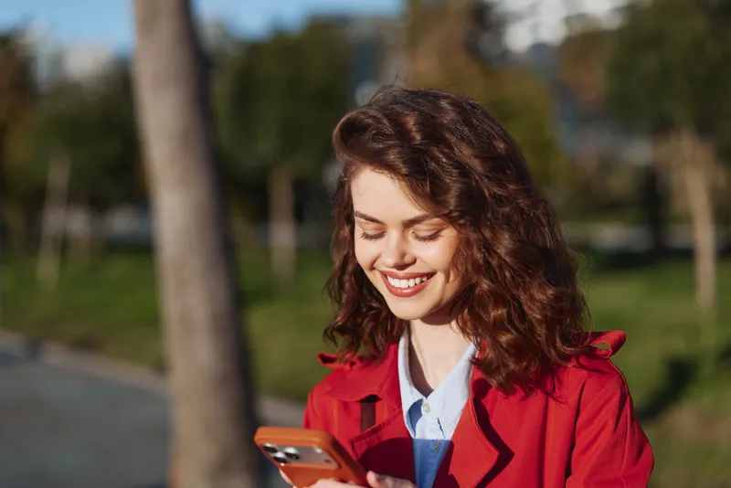 Bright outdoor portrait of a smiling woman in a red jacket using a smartphone in a park on a sunny day, casual candid moment, relaxed lifestyle scene.