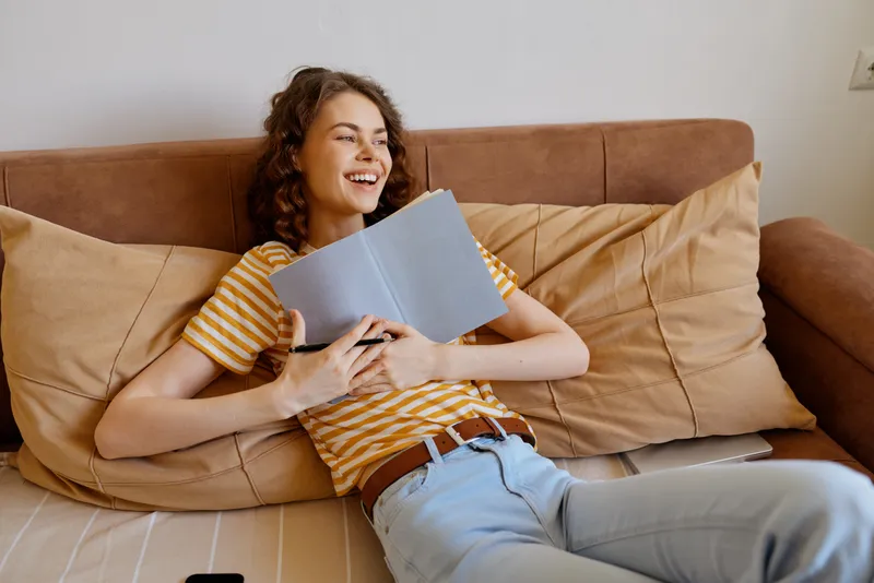 Woman relaxing on a couch at home, reading a book with a bright smile, casual striped top, jeans, cozy living room, warm light, lifestyle moment, enjoying quiet afternoon