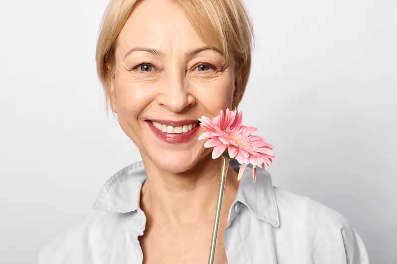 Smiling woman portrait in studio, blonde hair, holding a pink flower near her mouth, soft light, white background, close up, gentle focus headshot, cheerful mood, bright