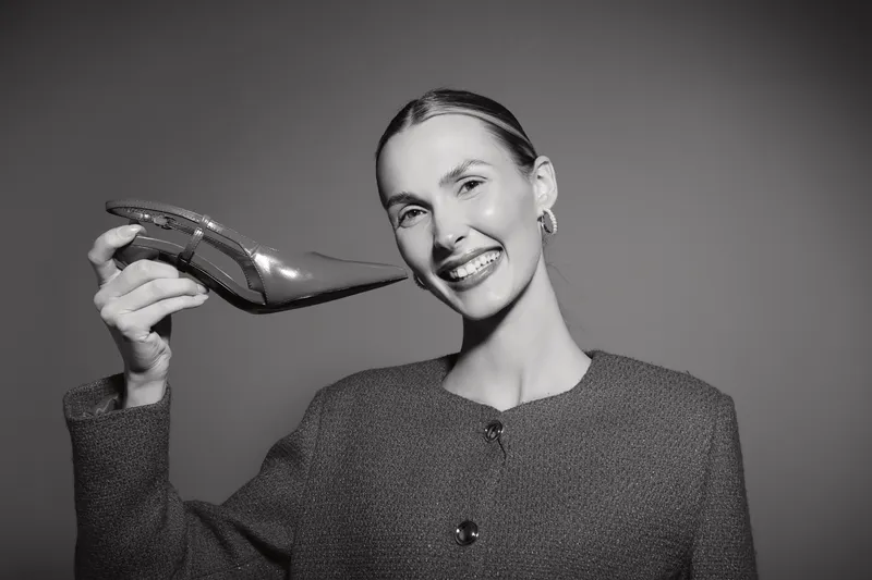 Smiling Caucasian woman with light skin and slicked-back hair holds a shiny shoe next to her ear. Studio portrait in black and white. Concept for fashion and happiness.