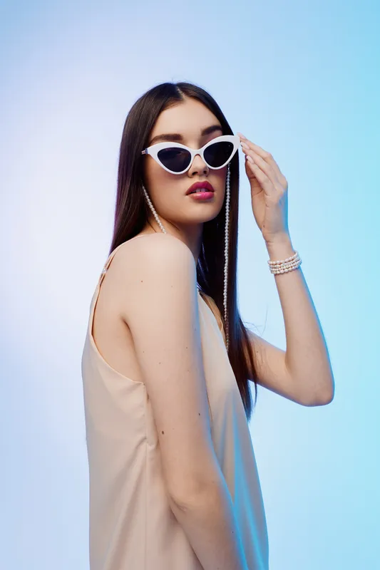 Young woman beauty fashion model wearing stylish white sunglasses and beige dress poses with confident expression on light blue background. Concept for modern style and elegance.