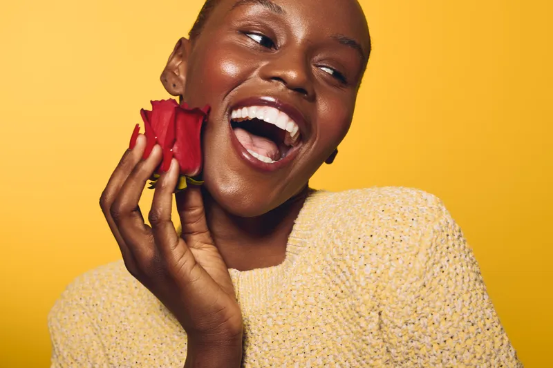 Happy African woman with dark skin laughs holding a red rose near her face. Yellow background and cozy beige sweater create warm, joyful, and vibrant mood for beauty and happiness concept.
