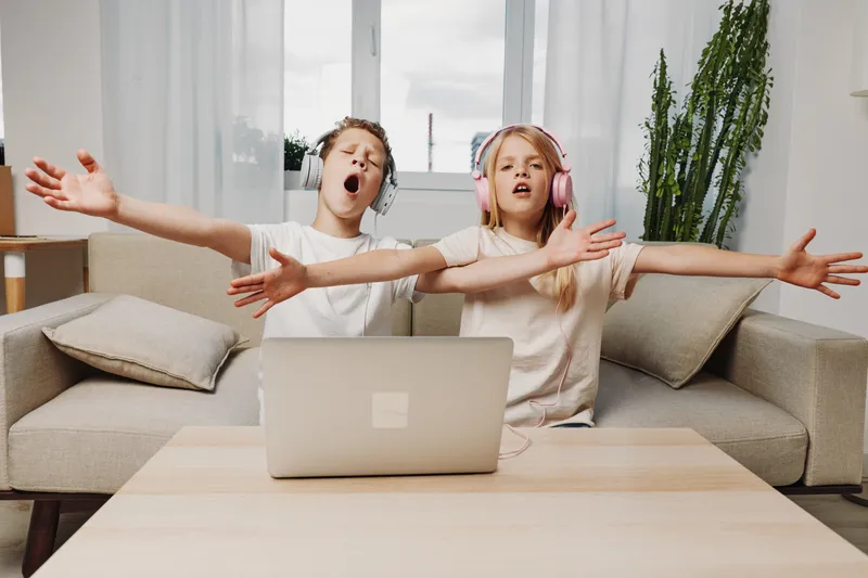 Two children wearing headphones sing passionately with arms wide open near a laptop on a wooden table in a bright living room. Concept of music enjoyment, childhood fun, and creativity.