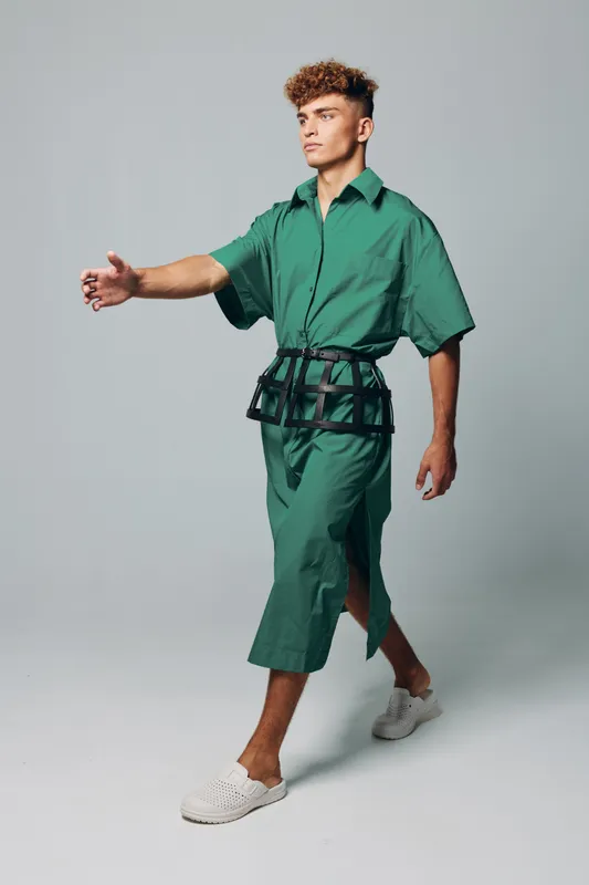Young man walking in green oversized shirt and matching wide pants with black belt accessory and white slip-on shoes on gray background in studio fashion portrait.