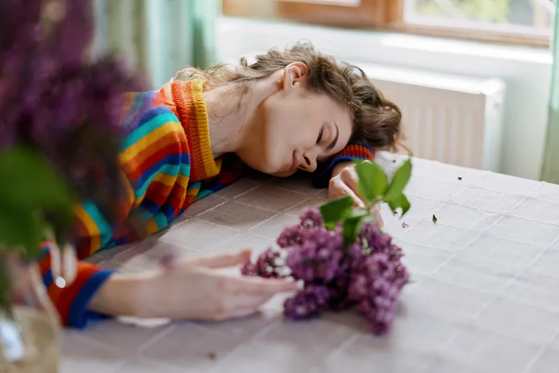 Sleeping woman with a colorful striped sweater rests her head on a table beside a purple bouquet, cozy home environment, calm mood, intimate domestic moment, soft light and quiet atmosphere.