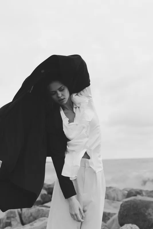 Monochrome fashion shot of a woman in a white dress with a dramatic black cloak by the sea, wind swept pose and minimalist elegance.