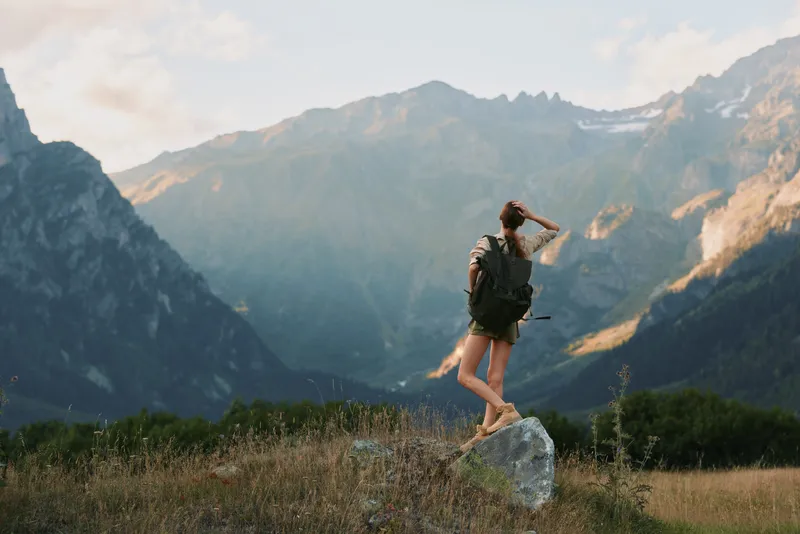 Young woman with backpack balances on a sunlit rock in a wide mountain meadow, enjoying a peaceful outdoor hike and fresh alpine air as distant peaks loom and a soft breeze