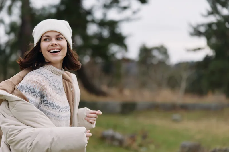 Woman outdoors in winter, smiling, wearing a jacket, sweater and beanie, standing in nature with countryside scenery and daylight, casual fashion mood and relaxed energy.