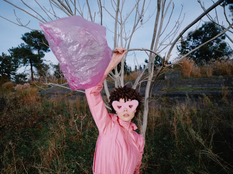 Pink woman outdoors wearing a pink scarf and mask, raising a translucent bag in a windy scene with bare trees, creating an artistic, surreal portrait.