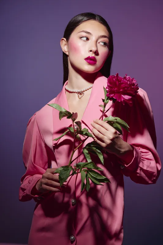 Young woman with glossy ruby lips and satin pink blouse holds a large vibrant flower indoors on a soft purple background. Concept of beauty creative fashion, elegance, and floral art.