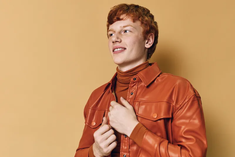 Young man with red hair wearing orange leather jacket and turtleneck sweater looking sideways with confident smile against light beige background in studio.