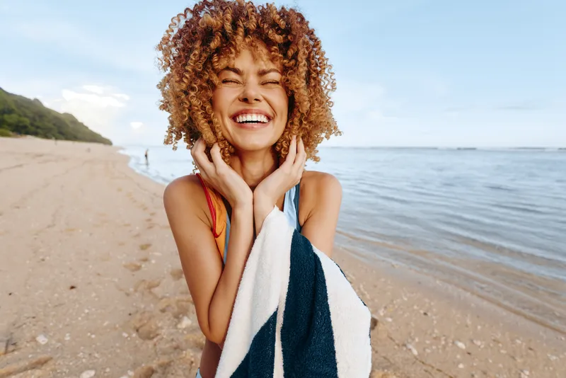 Happy young woman with curly hair wrapped in a striped towel enjoys a sunny day smiling on a sandy beach by the calm sea.