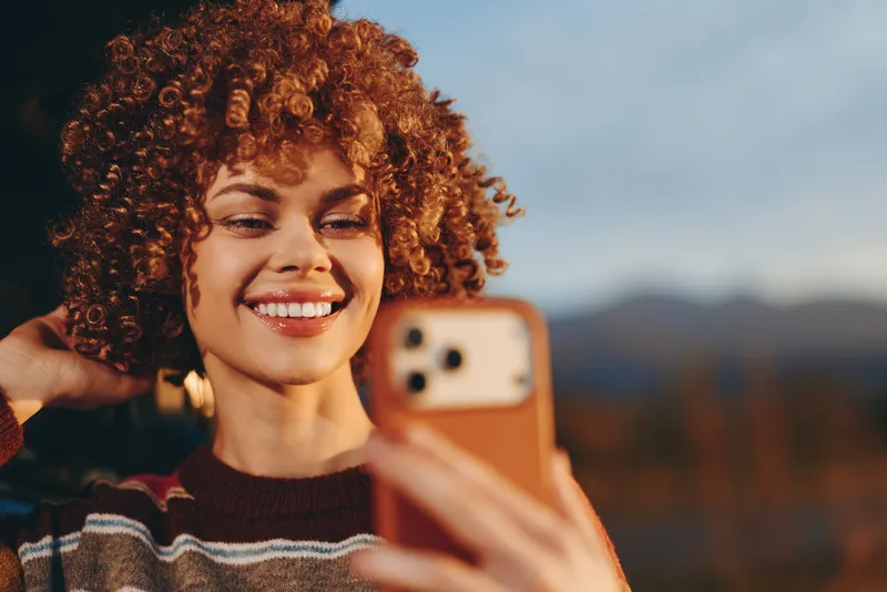 Smiling woman, curly hair, taking a selfie with a phone outdoors, holding the phone near her face, warm lifestyle moment with soft sunset light, blurred mountains bokeh background