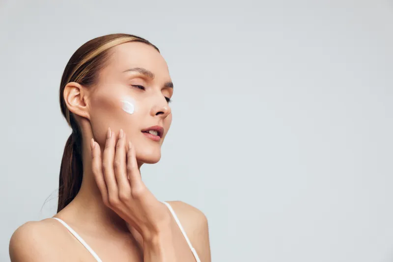 Young woman with a dab of cream on her cheek applies skincare in soft studio lighting, conveying a calm moment of self care and wellness, with a neutral background.