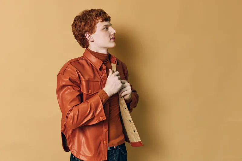 Young man with red hair wearing orange jacket holding his collar against beige wall looking sideways with confident expression in casual studio portrait photography.