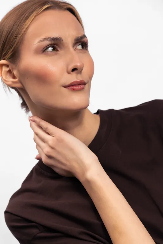 Young woman with short hair and clear skin looks thoughtfully to the side while touching her neck. Minimalist style with soft natural lighting on white background.