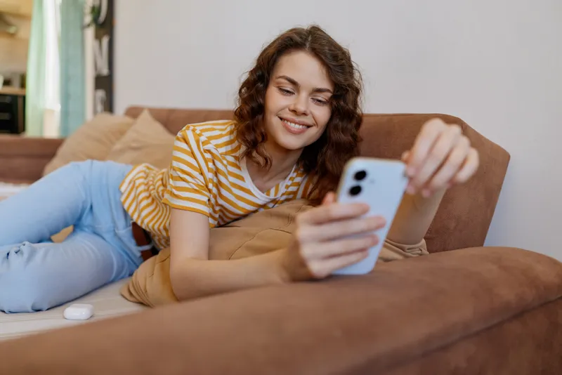 Young woman relaxes on a cozy living room sofa at home, smiling as she scrolls on a smartphone in a striped tee and light jeans.