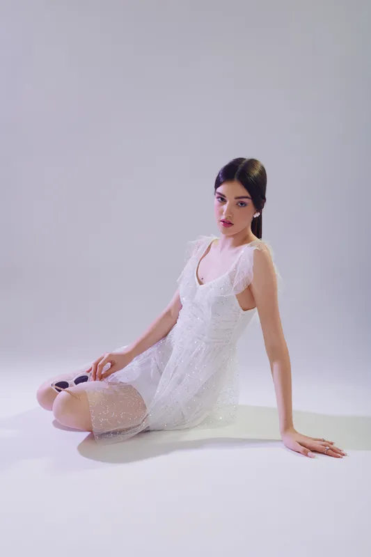 Young woman in a white lace dress sits on the floor against a plain background. Her dark hair is styled neatly. Concept of beauty, fashion, and elegance in studio lighting.