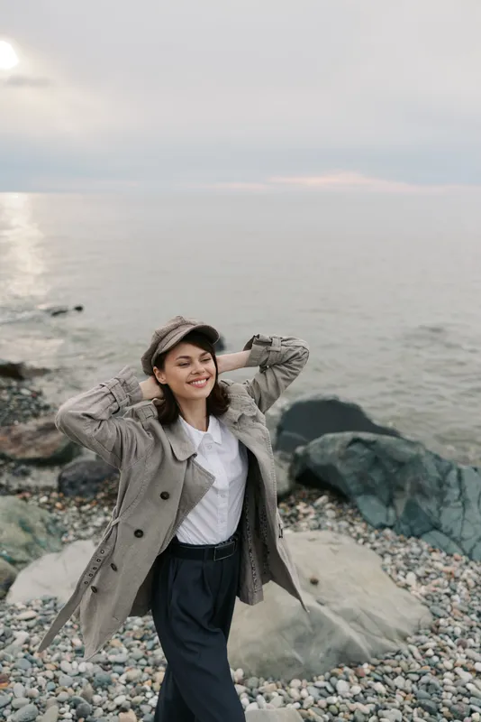 Smiling young woman in coat and cap enjoys sea breeze on rocky beach. Relaxed female model poses with hands behind head by calm water under cloudy sky.