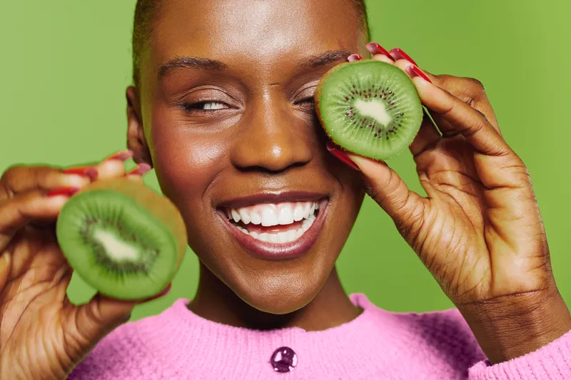 Happy African woman with dark skin holds fresh kiwi slices near face smiling with closed eyes on green background. Concept of healthy lifestyle, natural food, and joyful wellness.