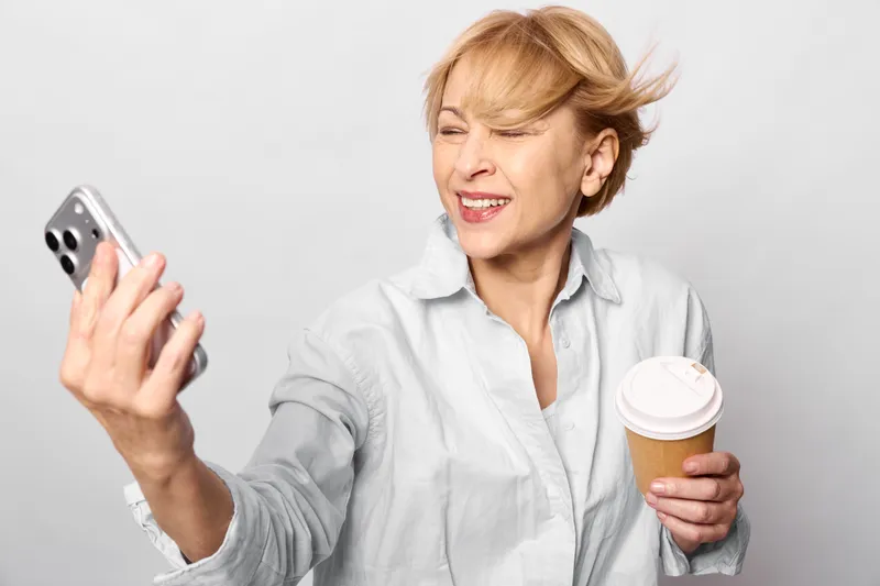 Woman uses smartphone, holding coffee mug, older, on gray background, expressive frustration emotion, communication, indoor casual moment, beverage break, headshot, closeup
